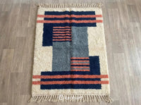 Custom handmade Beni Ourain Moroccan rug flat lay showing plush natural cream wool texture with dark blue, blue grey, and blue coral geometric diamond patterns and traditional fringe.